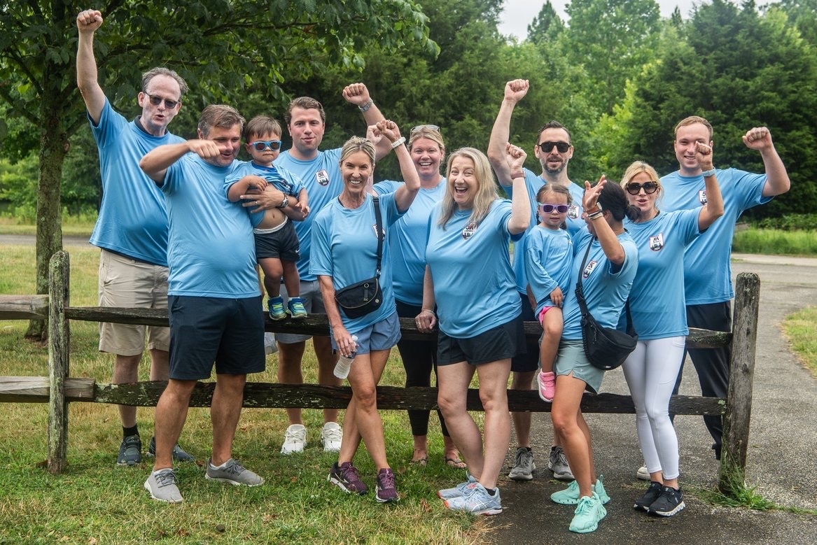 Berkadia team members in matching blue shirts celebrating together at the Coaches vs. Cancer 5K with raised fists at Wilson Farm Park